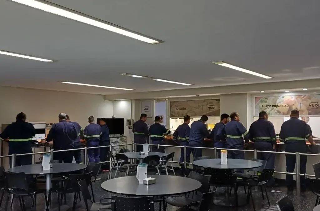 Colaboradores en la línea de servicio de un comedor corporativo con mesas disponibles en el salón.